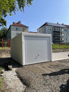Eine weiße Garage steht auf einem Kiesplatz mit Wohnhäusern und einem klaren blauen Himmel im Hintergrund.