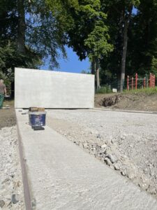 Eine große Betonwandplatte liegt an einem sonnigen Tag auf einer von Bäumen und Kies umgebenen Baustelle.