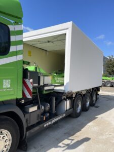 Grüner und weißer Lastwagen mit offener weißer Ladekiste, in der grüne Maschinen zu sehen sind, der an einem sonnigen Tag im Freien geparkt ist.