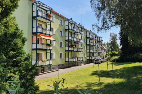 Ein grünes Mehrfamilienhaus mit Balkonen, Pflanzen und einem Rasen davor, Bäumen und geparkten Autos daneben.